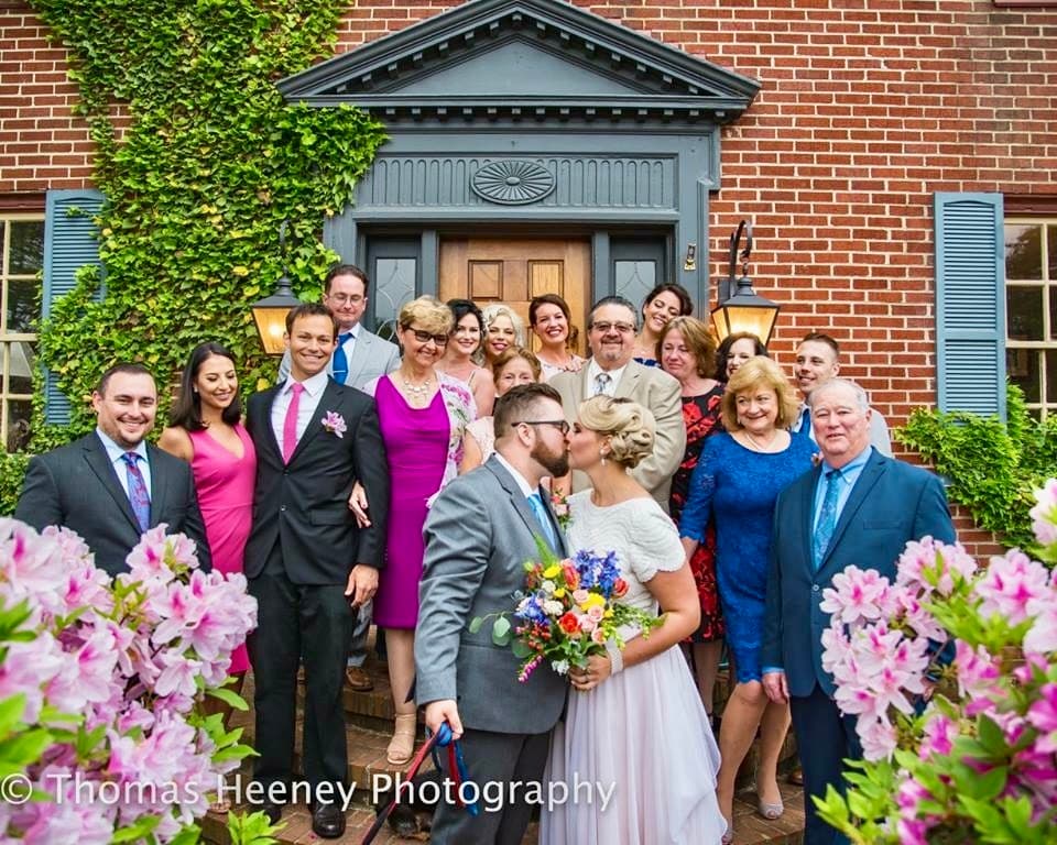 A wedding party poses together in front of a charming brick house, with the bride and groom kissing in the foreground surrounded by flowers.