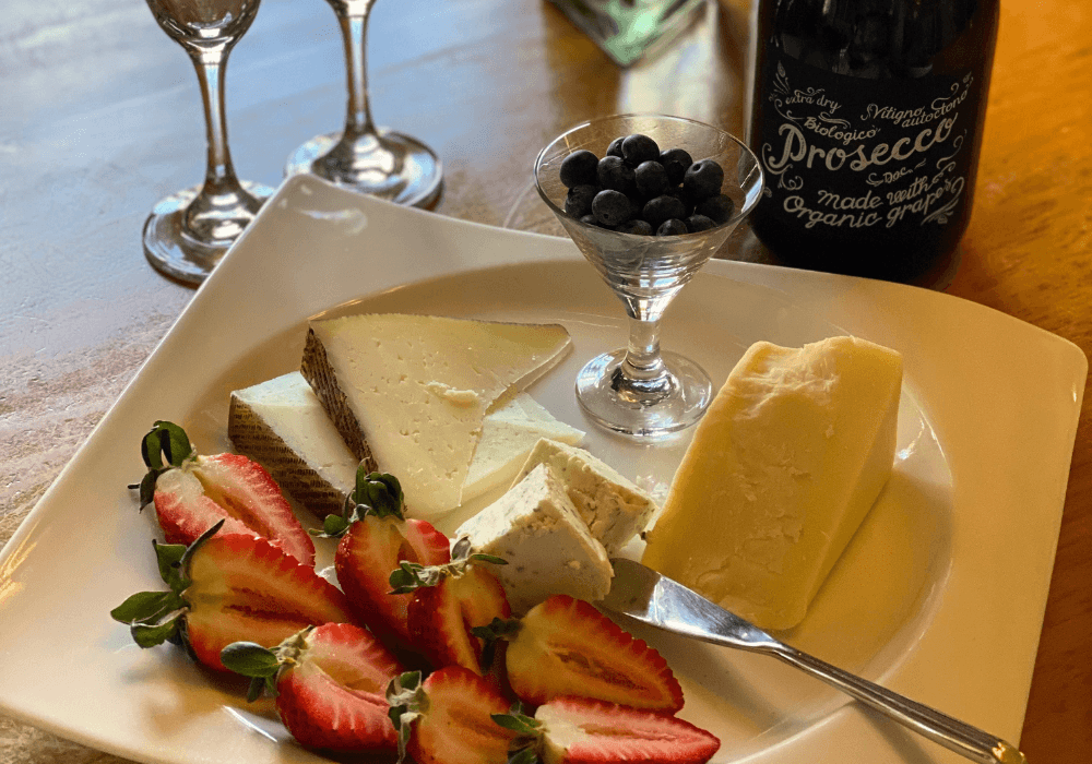 A platter of assorted cheeses, fresh strawberries, and blueberries, accompanied by a bottle of Prosecco and two wine glasses.