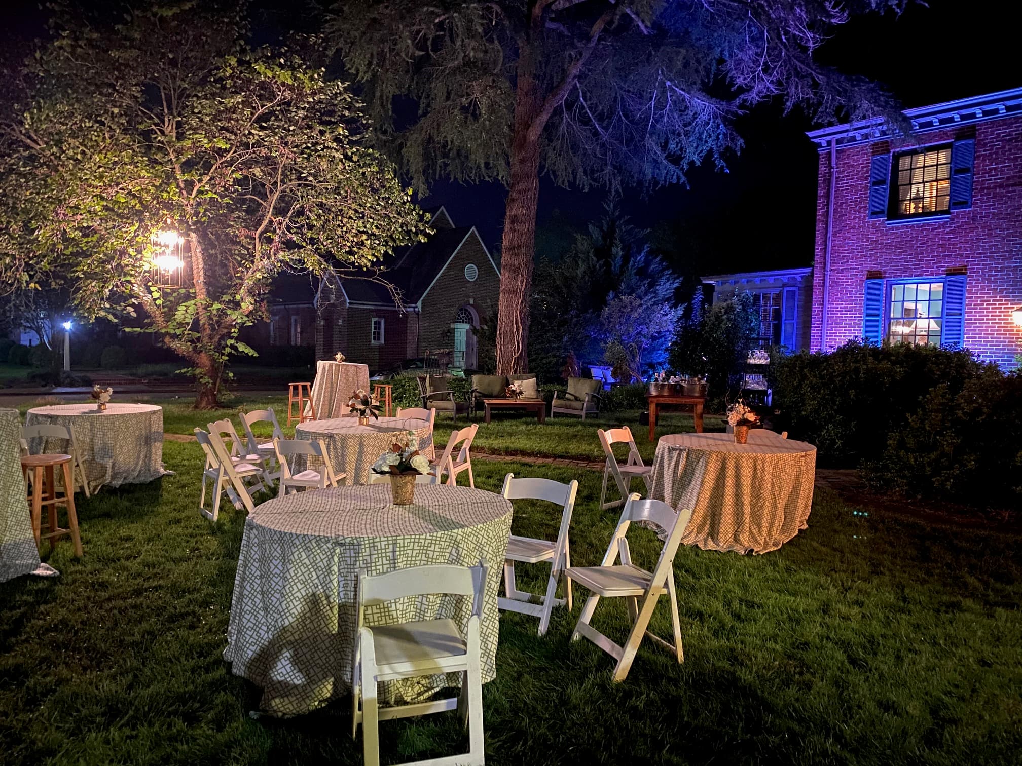 An outdoor event space at night featuring several decorated tables and chairs surrounded by trees and illuminated by soft lighting.