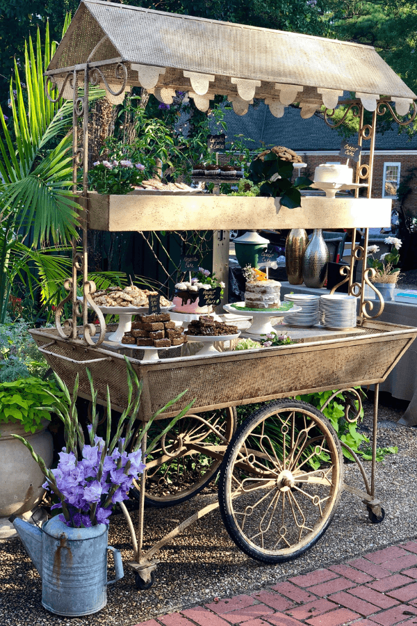 A vintage dessert cart displays an array of cakes, cookies, and pastries, adorned with greenery and flowers.