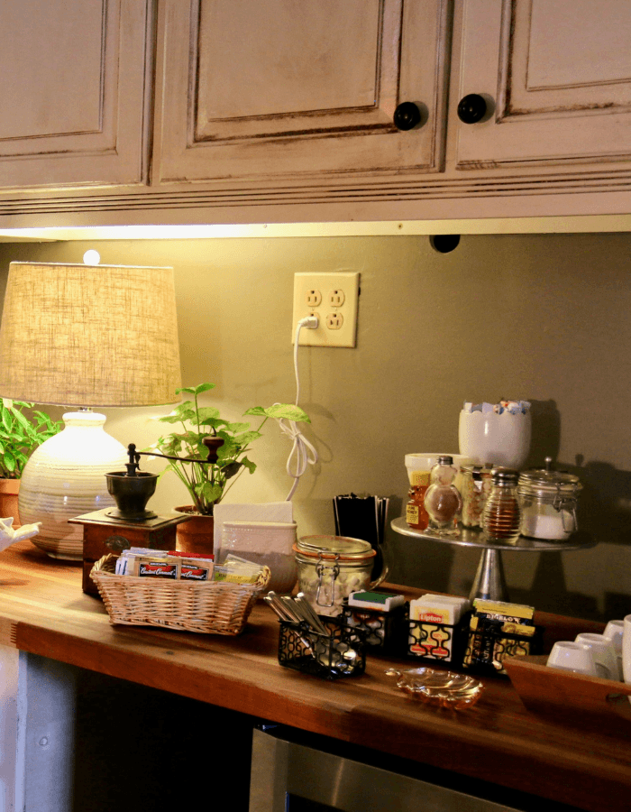 A cozy kitchen counter featuring a lamp, plants, various condiments, and tea supplies.