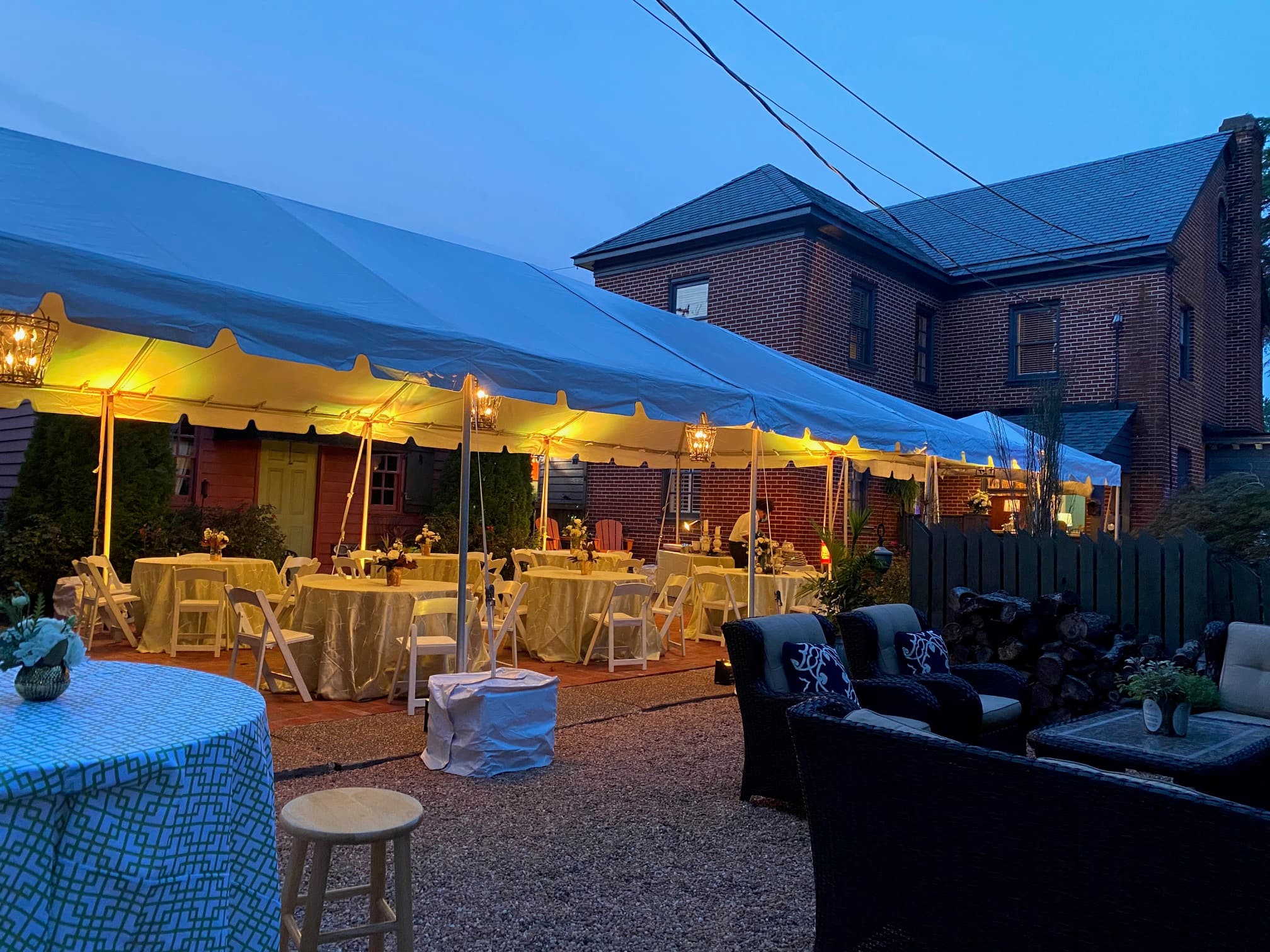 A backyard event set up under a large tent, featuring tables and chairs, illuminated in the evening light.