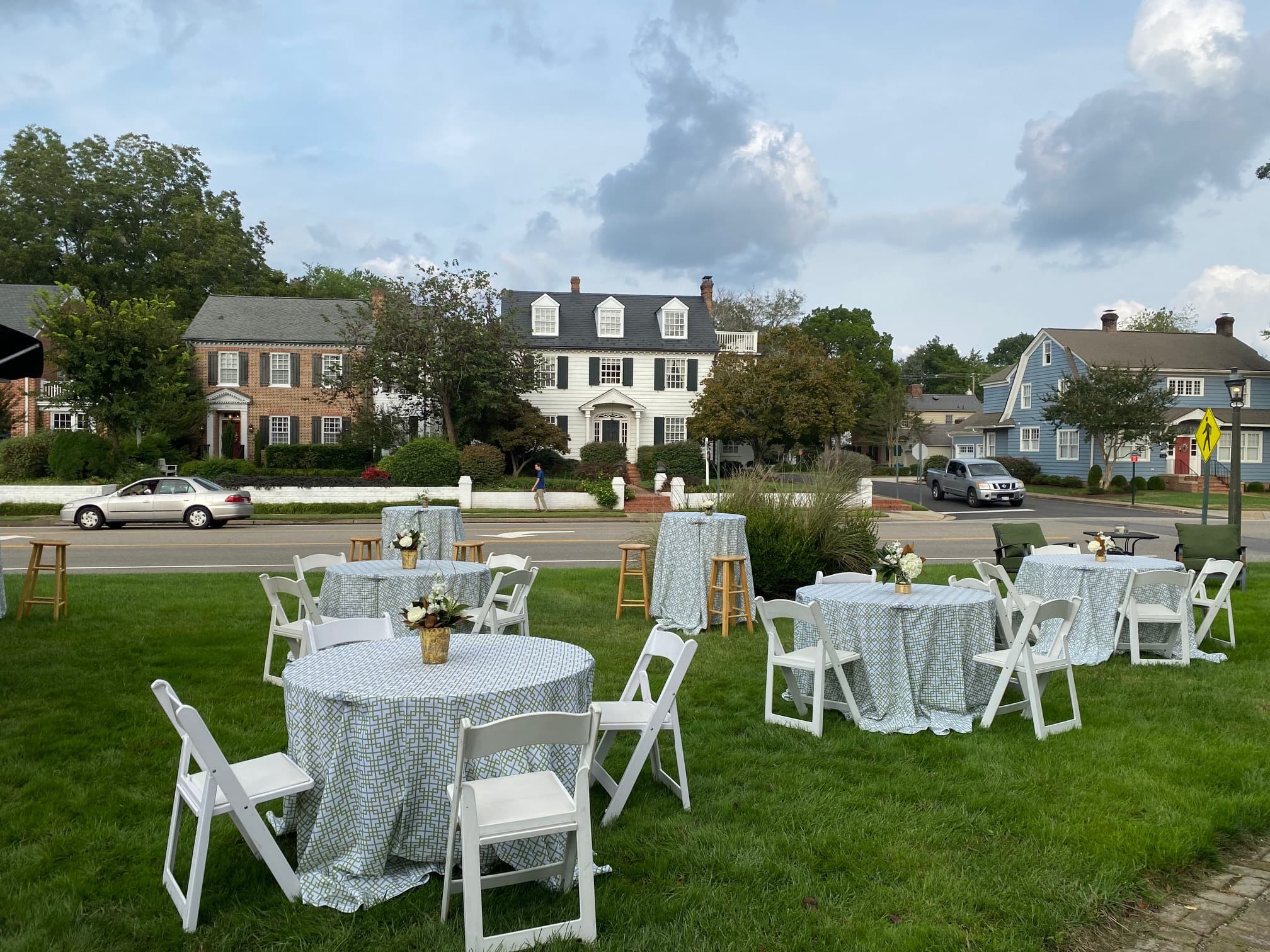 Outdoor event setup featuring round tables with floral centerpieces on a grassy lawn, surrounded by houses and a cloudy sky.