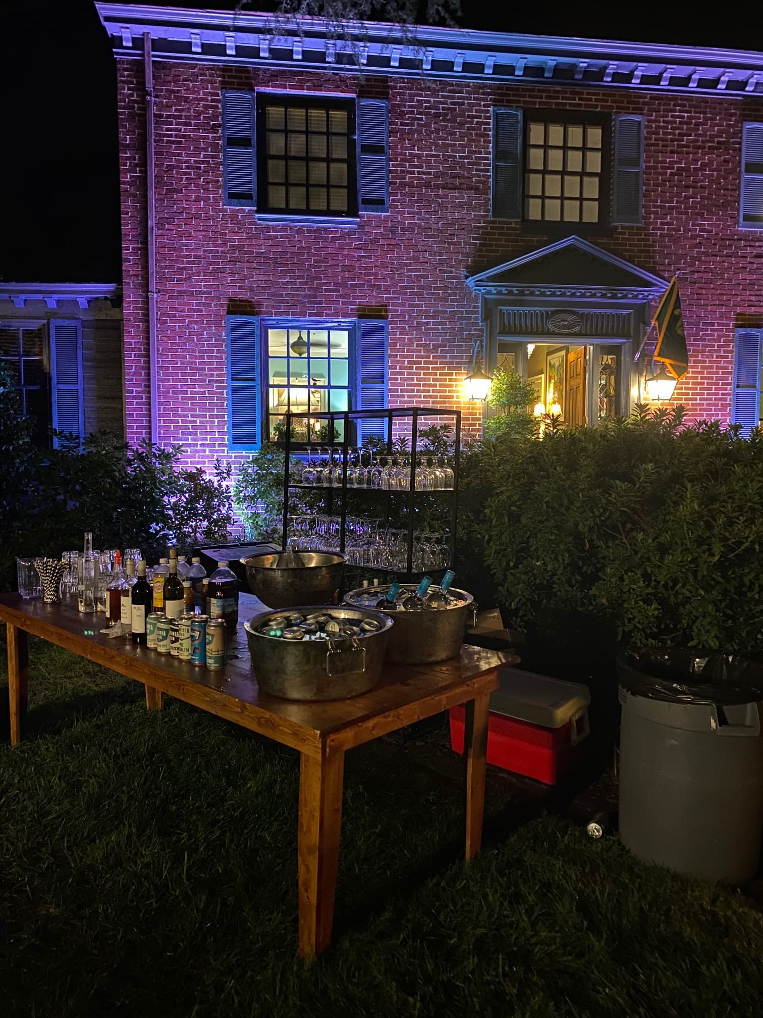 A well-lit outdoor bar setup with drinks in front of a charming, illuminated brick house at night.