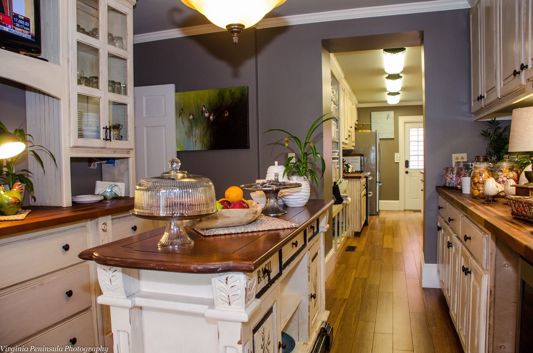 A warmly lit kitchen features wooden cabinetry, a central island with a glass dome, and a view of a doorway leading to an exterior entrance.