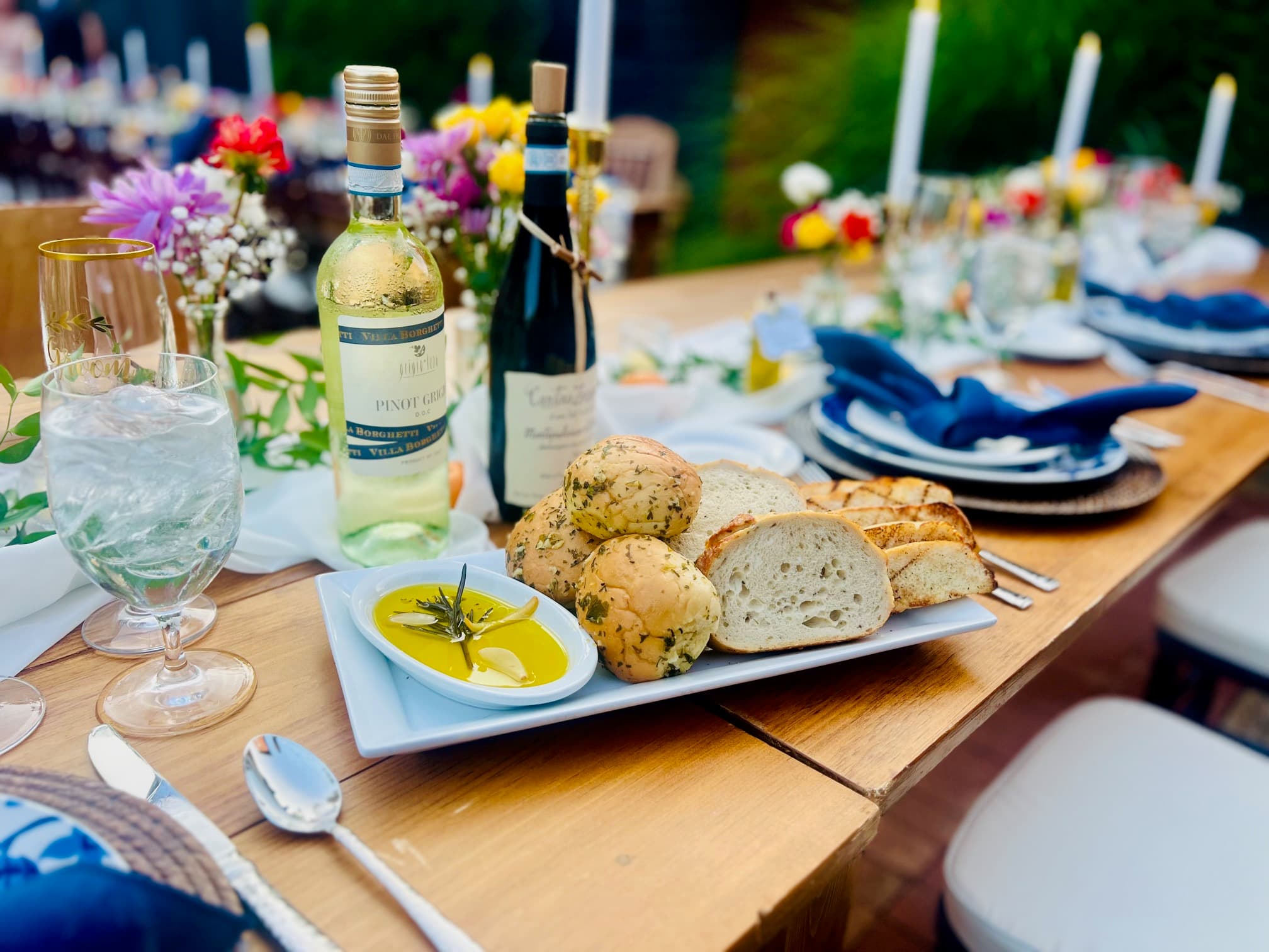 A beautifully set dining table featuring a platter of assorted breads, olive oil, and two bottles of wine surrounded by floral centerpieces and candlelight.