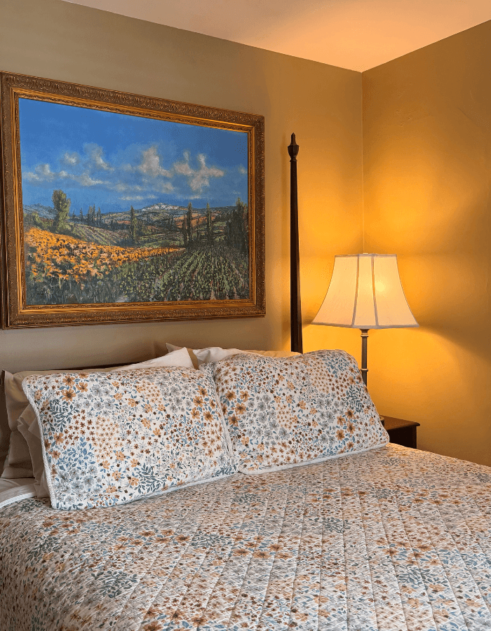 A cozy bedroom featuring a floral-patterned bedspread, decorative pillows, a bedside lamp, and a landscape painting on the wall.