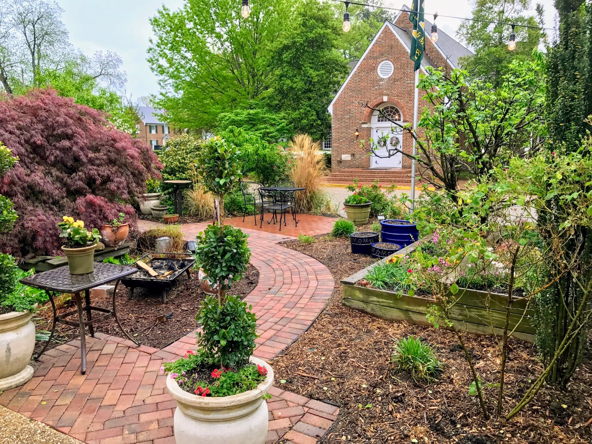 A winding brick path surrounded by vibrant greenery and potted plants leads to a rustic building in a serene garden setting.