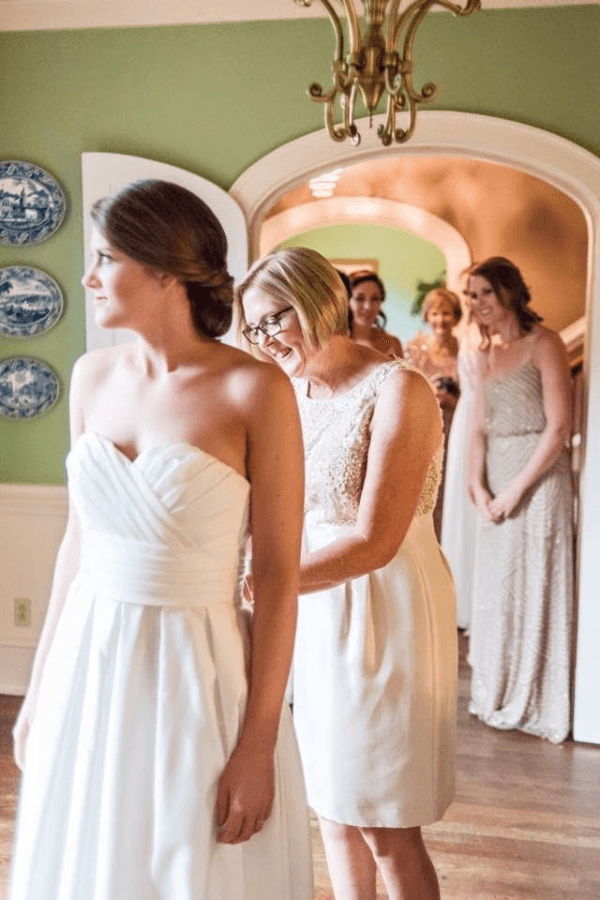 A bride is getting dressed with the assistance of her mother while other bridesmaids look on in the background.