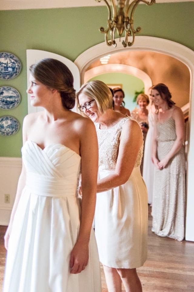 A bride stands in a white gown while her mother adjusts it, surrounded by bridesmaids in the background.