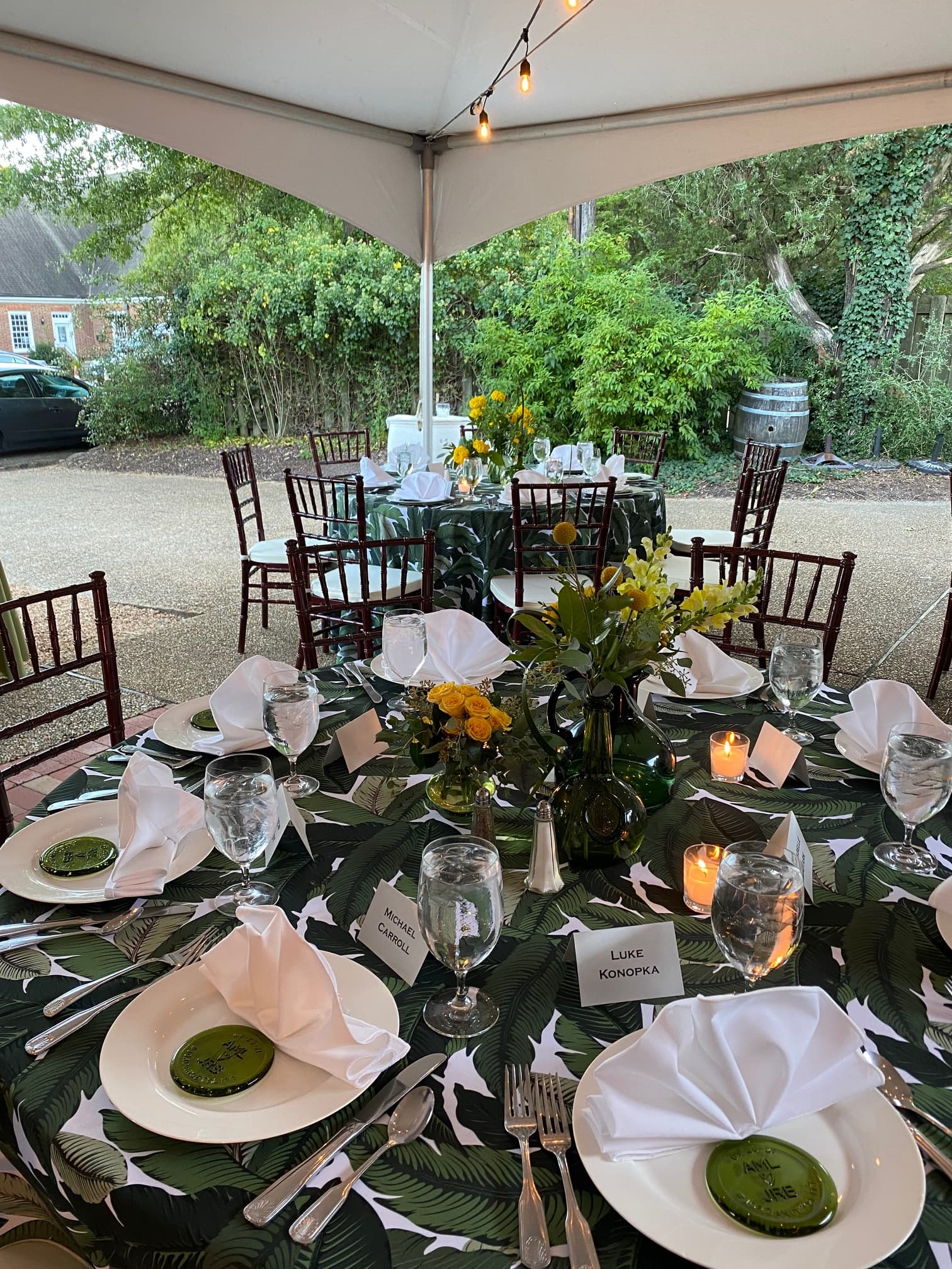 A beautifully assembled outdoor dining table with a leafy tablecloth, flower centerpieces, and place cards under a tent.
