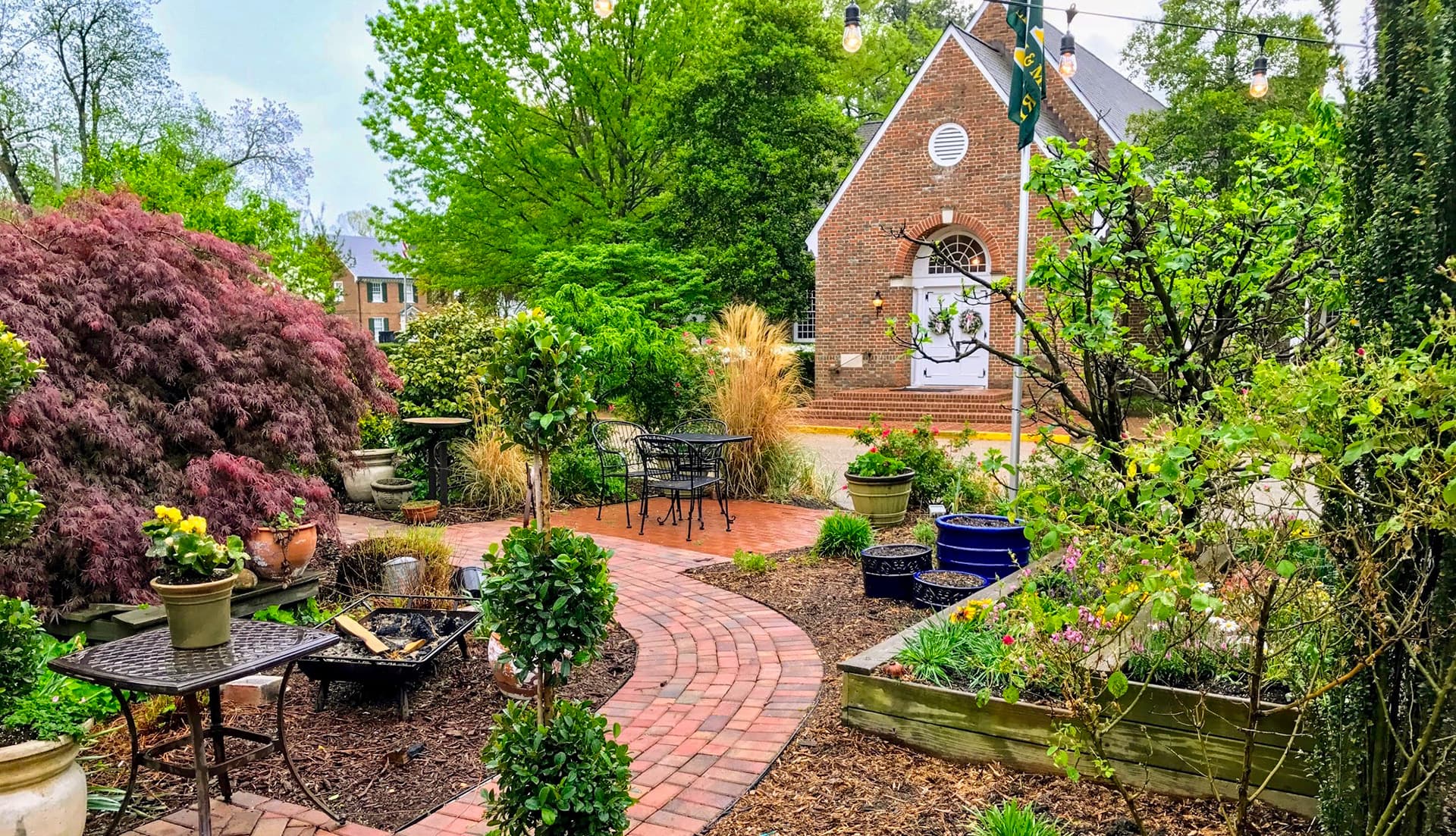 A peaceful garden pathway winding through lush greenery and potted plants, leading to a brick building in the background.