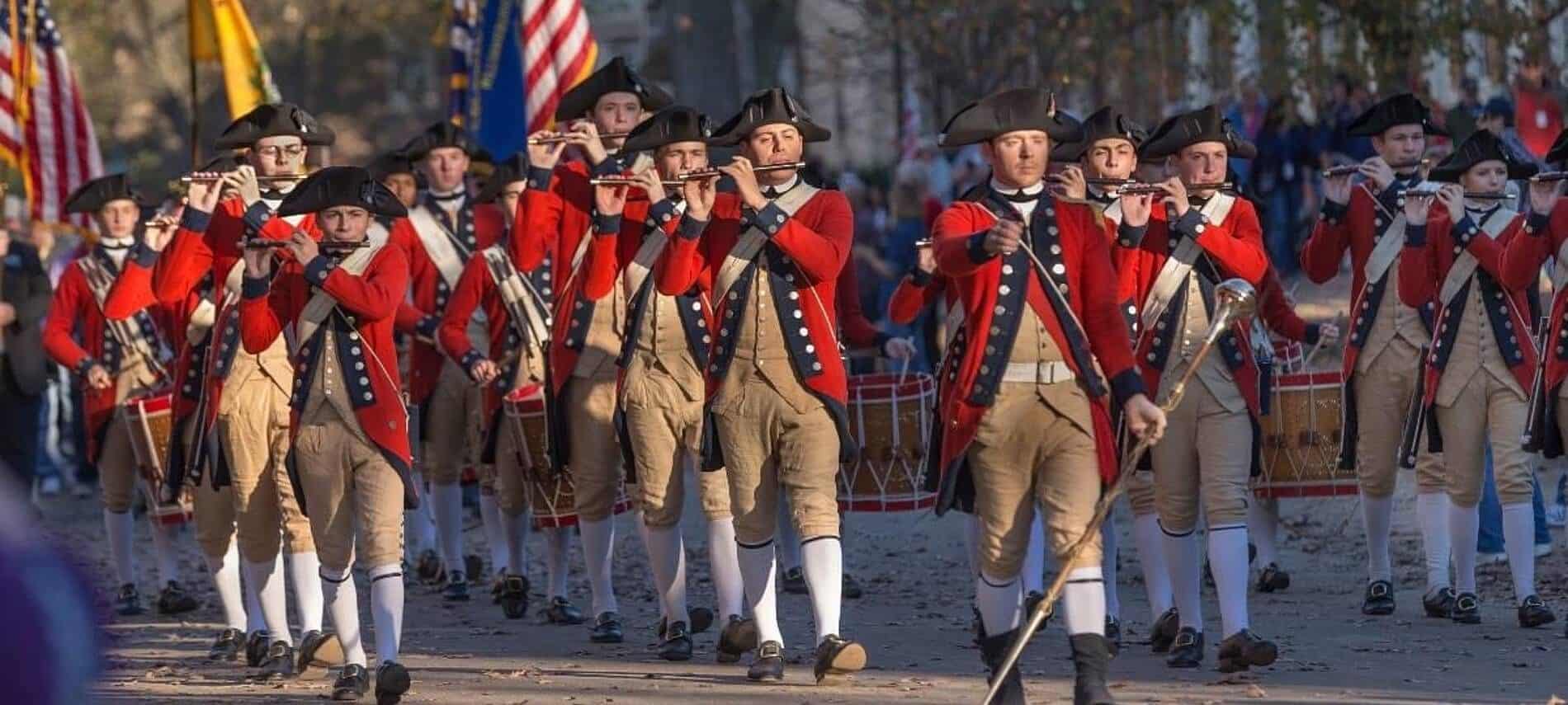 Large group of men dressed in colonial costumes and playing flutes marching down a street