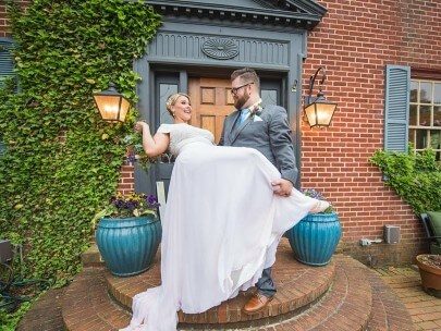 Smiling bride in white dress being dipped by her groom in a grey suit in front of a brick home