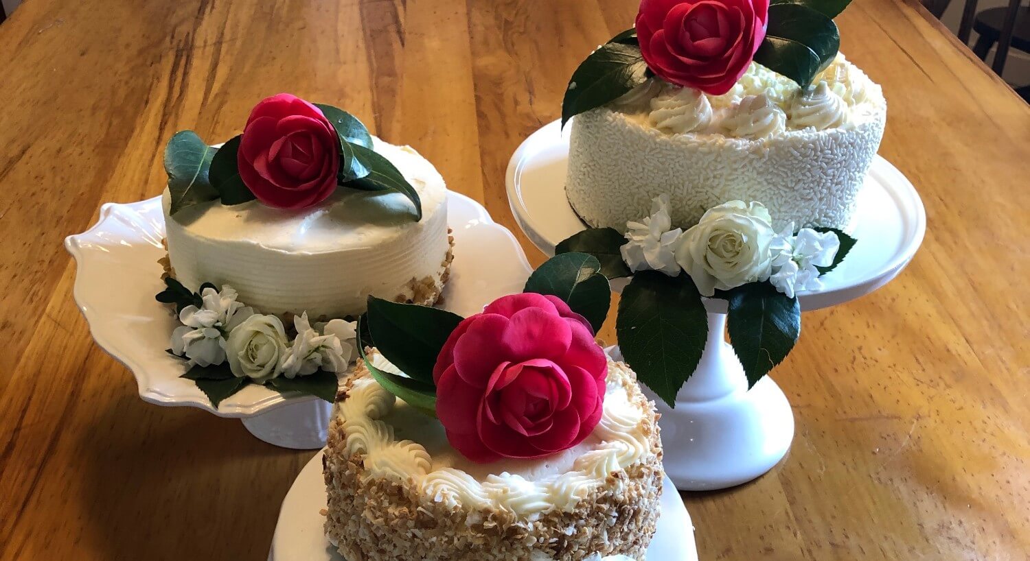 Three small white cakes on white stands decorated with red and white flowers and green leaves