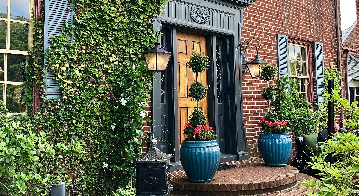Front door of large brick home with black trim, rounded steps and large blue planters with red flowers and topiary trees