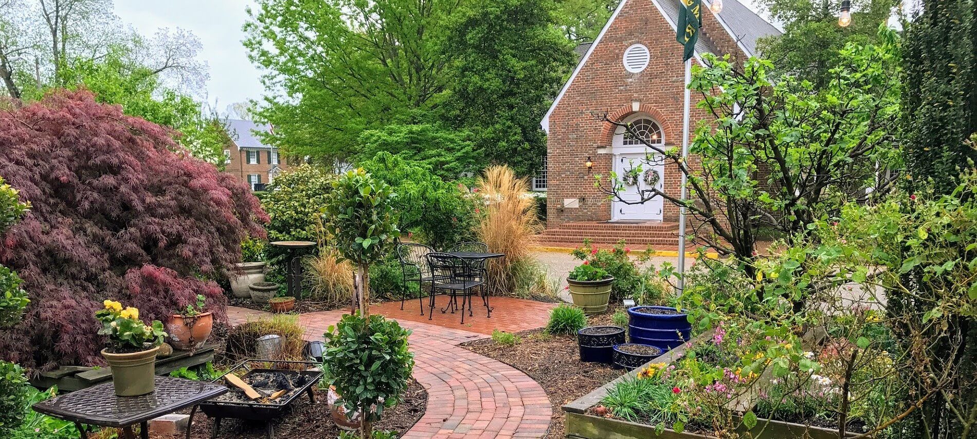 Outdoor garden patio with several planters, two black tables, brick path and tall brick building in background