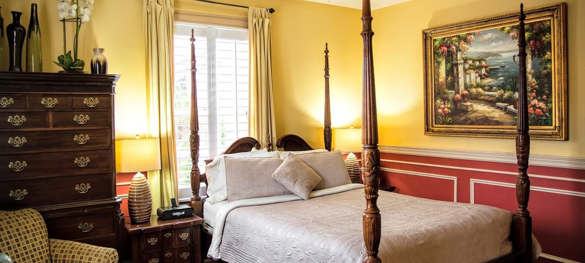 Elegant guest room with four poster bed in white linens, red and yellow walls and brown accent furniture