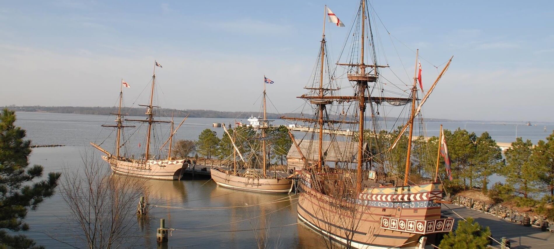 Harbor area showing three wooden sailing ships docked next to a pathway lined with trees