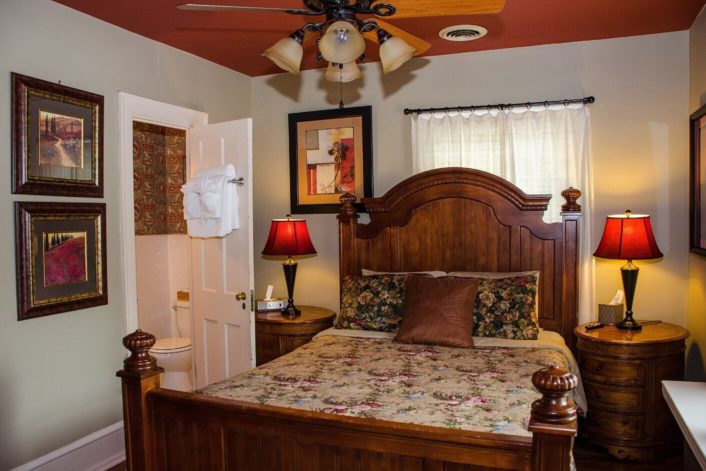 Guest room featuring queen bed with tall brown headboard with window behind, round side tables and doorway into bathroom