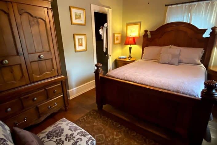 Elegant guest room with brown queen bed, large armoire, sitting chair and doorway into small bathroom