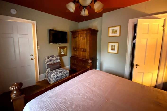 Guest room featuring queen bed in white linens, brown armoire next to sitting chair and TV on the wall