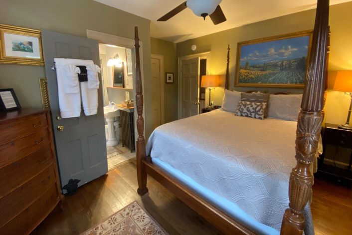 Cozy guest room with four poster bed in white bedding, brown dresser and side tables and doorway into small bathroom