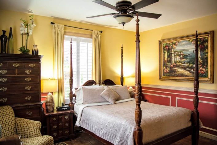 Four poster bed in guest room with red and yellow walls, dark brown dresser and side table, and upholstered chair