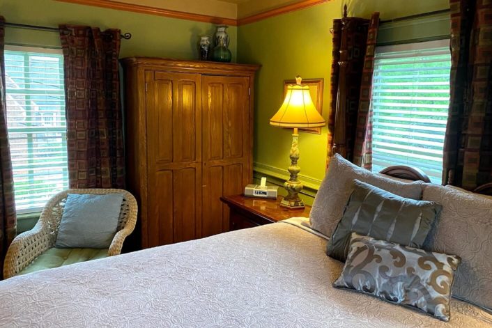 Cozy guestroom with grey bedding and pillows, tall brown armoire, chair, two windows and side table with lamp