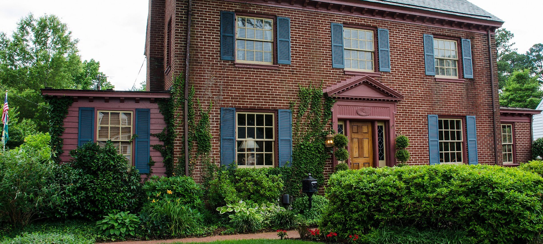 Facade of a tall red brick home with windows flanked by blue shutters, brown front door and lush green shrubs