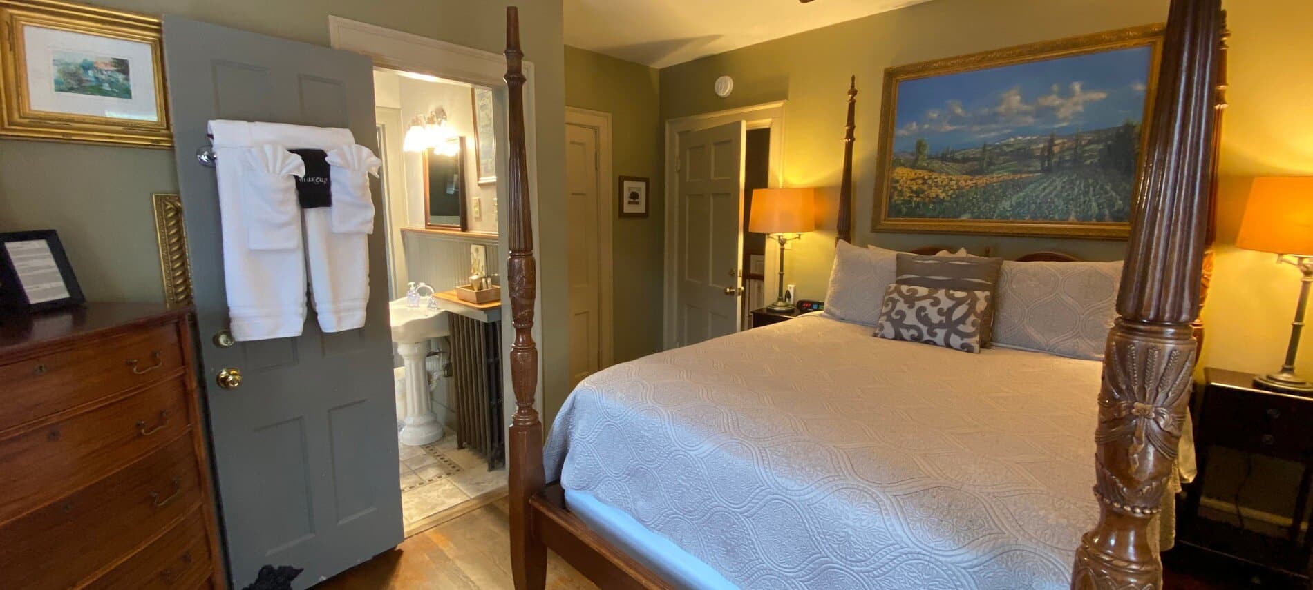 Elegant guest room with beautiful four poster bed in white linens, open doorway into a bathroom and brown dresser in corner