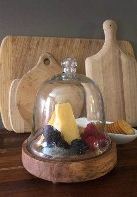 Wooden cutting boards of various sizes behind a glass cloche of fruits and cheese and platter of crackers