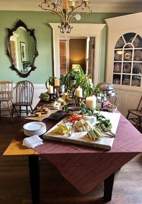 Dining table withe chairs pushed aside featuring a buffet of a variety of foods and tall white candles