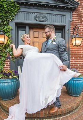 Groom in grey suit dipping a smiling bride in white at the doorway of a red brick home