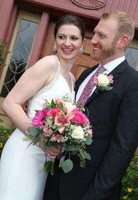 Smiling bride holding a pink and white bouquet next to her groom in black in front of a door outside