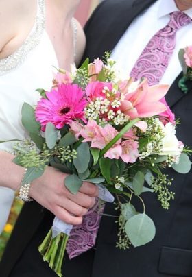 Groom in black suit next to a bride in white holding a flower bouquet of pink and white flowers