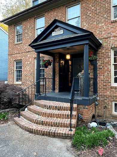 Front porch of a brick home with black peaked overhang, curved steps and hanging baskets by door